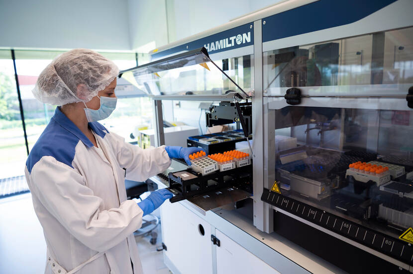 Onderzoekster draagt een labjas, haarnetje en handschoenen en zet bakjes gevuld met buisjes in een apparaat.