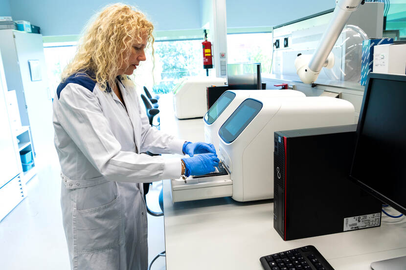 Onderzoekster in een labjas en met handschoenen aan bekijkt een analyse-apparaat op het lab.
