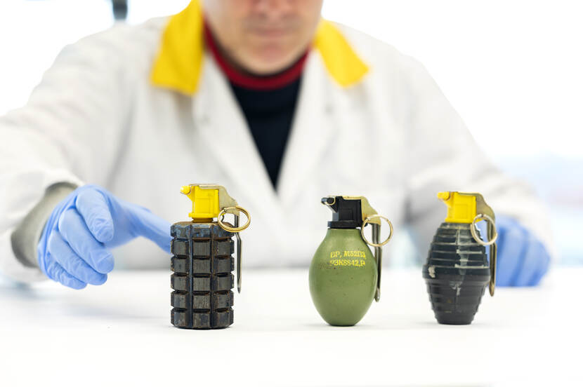 Drie verschillende handgranaten staan op een labtafel terwijl een van de onderzoekers met zijn hand er naar reikt.