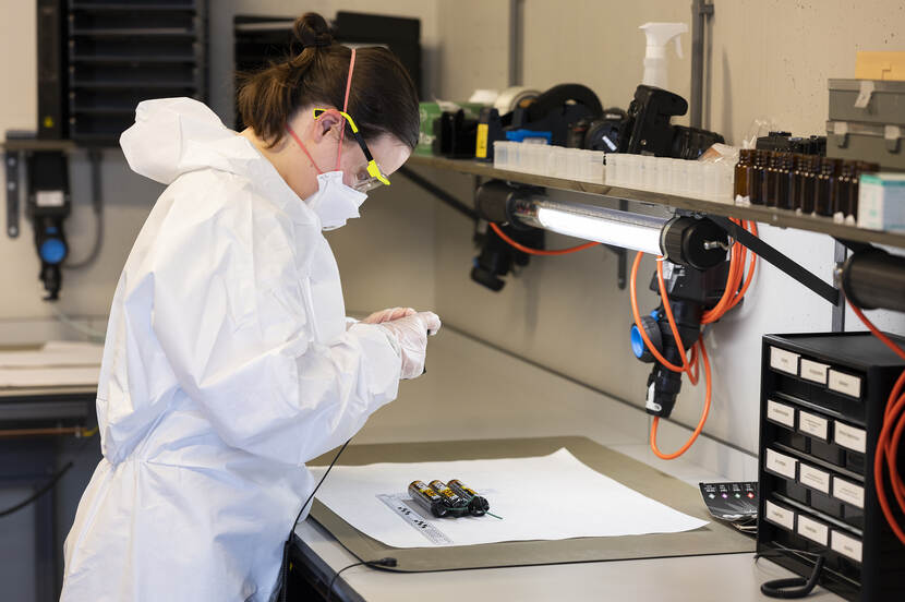 Onderzoeker in een witte overall met mondkapje en beschermingsbril maakt een foto terwijl een stukken vuurwerk op een onderzoekstafel ligt.